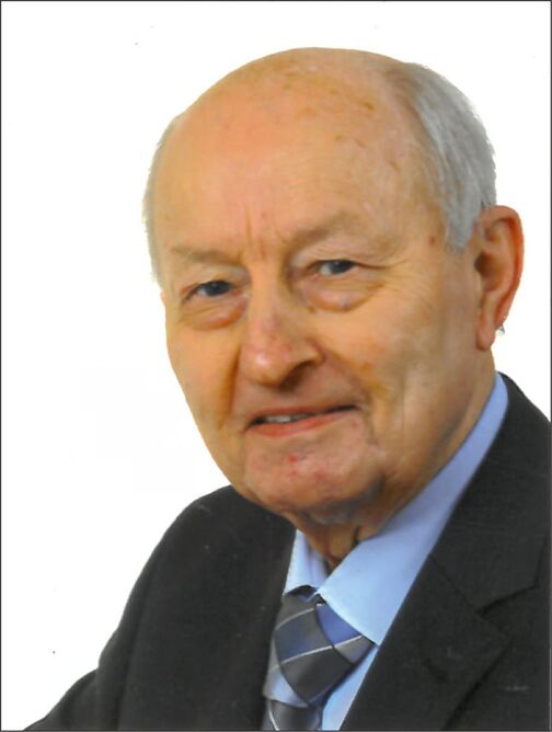 Portr&auml;t eines &auml;lteren Mannes mit grauem Haar, in einem dunklen Anzug und blauem Hemd, l&auml;chelnd vor wei&szlig;em Hintergrund.