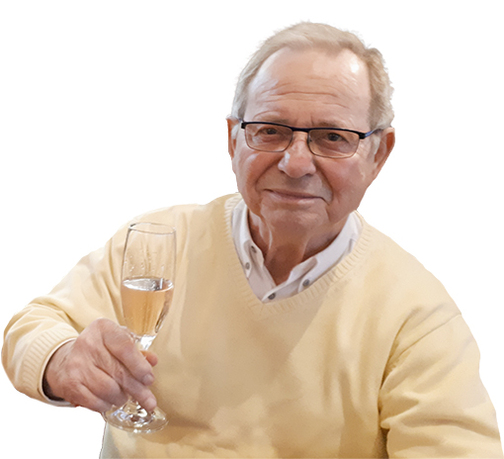 &Auml;lterer Mann in einem hellen Pullover h&auml;lt ein Sektglas und l&auml;chelt in die Kamera.