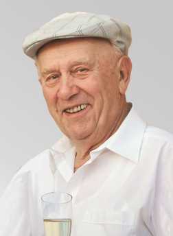 &Auml;lterer Mann mit einem L&auml;cheln, tr&auml;gt eine graue M&uuml;tze und ein wei&szlig;es Hemd, h&auml;lt ein Glas mit einem klaren Getr&auml;nk.