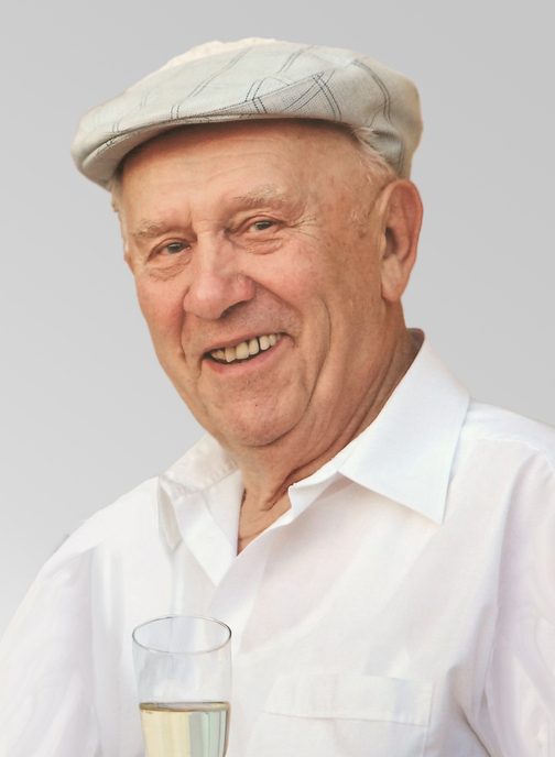 &Auml;lterer Mann mit einem L&auml;cheln, tr&auml;gt eine graue M&uuml;tze und ein wei&szlig;es Hemd, h&auml;lt ein Glas mit einem klaren Getr&auml;nk.