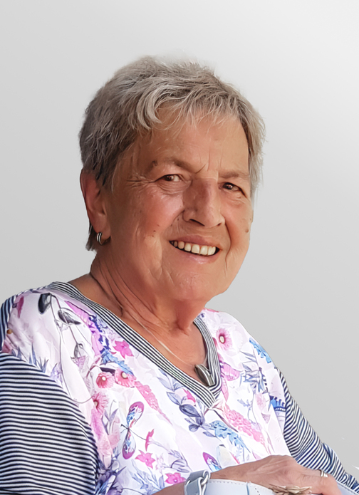 L&auml;chelnde &auml;ltere Frau mit kurzem grauen Haar, tr&auml;gt ein bl&uuml;hendes Oberteil und sitzt vor einem hellen Hintergrund.