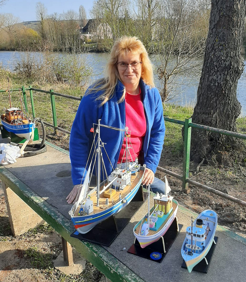 Frau mit blauer Jacke und Brille posiert mit mehreren Schiffsmodellen auf einem Tisch am Ufer eines Gew&auml;ssers.
