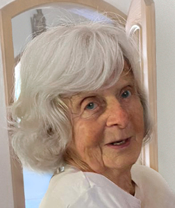 Portr&auml;t einer &auml;lteren Frau mit grauen Haaren, die in einen Raum schaut, mit einer Holzrahmen-T&uuml;r im Hintergrund.