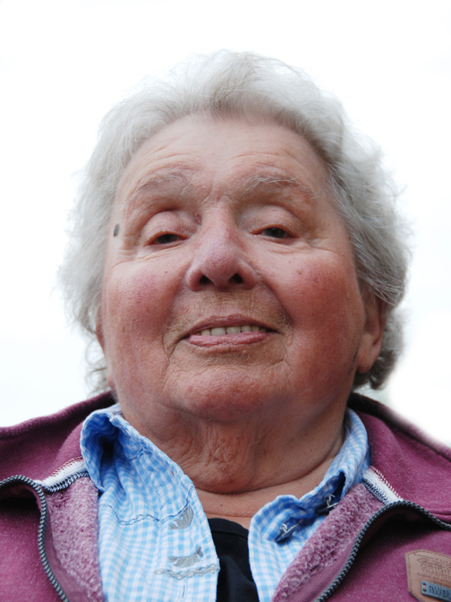 Nahaufnahme einer &auml;lteren Frau mit grauen Haaren, die freundlich in die Kamera l&auml;chelt. Sie tr&auml;gt ein kariertes Hemd und eine Jacke.