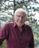 &Auml;lterer Mann mit Brille und kariertem Hemd steht vor einer Waldlandschaft mit B&auml;umen und gr&uuml;nen Wiesen im Hintergrund.