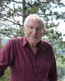 Älterer Mann mit Brille und kariertem Hemd steht vor einer Waldlandschaft mit Bäumen und grünen Wiesen im Hintergrund.