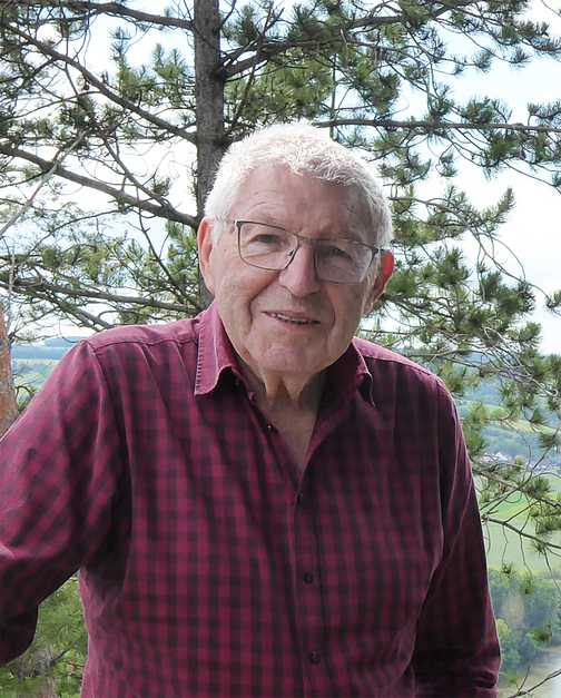 &Auml;lterer Mann mit Brille und kariertem Hemd steht vor einer Waldlandschaft mit B&auml;umen und gr&uuml;nen Wiesen im Hintergrund.