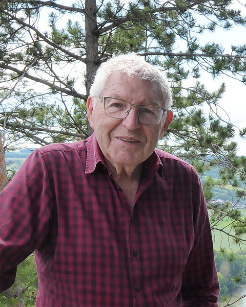 &Auml;lterer Mann mit Brille und kariertem Hemd steht vor einer Waldlandschaft mit B&auml;umen und gr&uuml;nen Wiesen im Hintergrund.