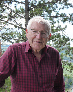 &Auml;lterer Mann mit Brille und kariertem Hemd steht vor einer Waldlandschaft mit B&auml;umen und gr&uuml;nen Wiesen im Hintergrund.