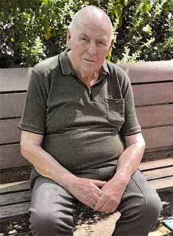 &Auml;lterer Mann sitzt auf einer Holzbank, tr&auml;gt ein graues Polohemd und blickt in die Kamera, umgeben von gr&uuml;nem Laub.