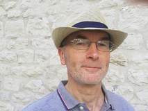Mann mit Hut und Brille vor einer hellen Steinmauer, l&auml;chelt sanft in die Kamera.