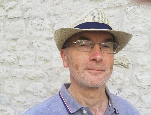 Mann mit Hut und Brille vor einer hellen Steinmauer, l&auml;chelt sanft in die Kamera.
