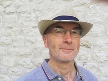 Mann mit Hut und Brille vor einer hellen Steinmauer, lächelt sanft in die Kamera.