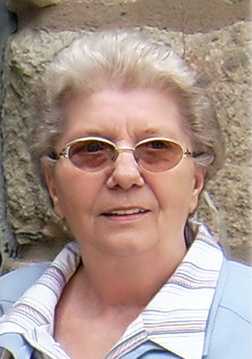 &Auml;ltere Frau mit Brille und blonder Haarfarbe, vor einer steinernen Wand stehend.