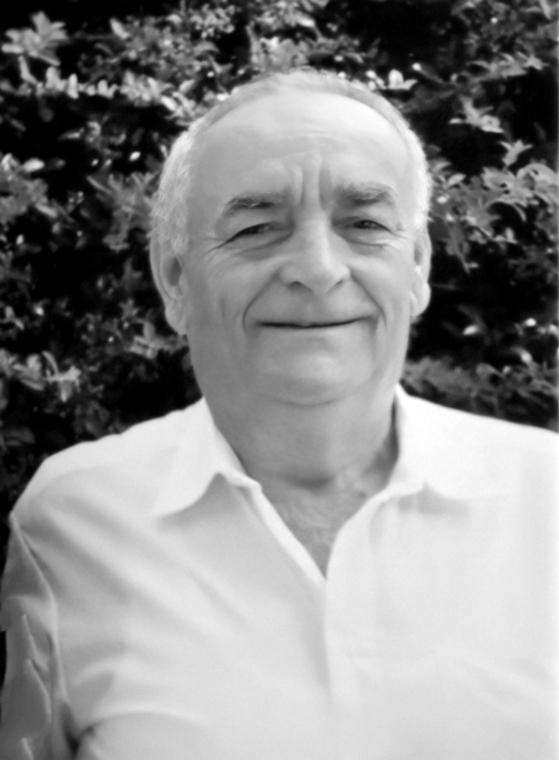 Schwarz-wei&szlig;-Portr&auml;t eines l&auml;chelnden Mannes mit kurzem, grauem Haar vor einer gr&uuml;nen Pflanze.