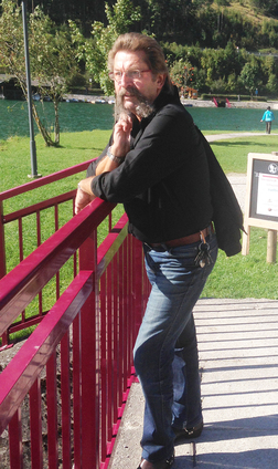 Mann mit Bart lehnt an einer roten Br&uuml;stung und blickt nachdenklich auf eine gr&uuml;ne Wiese und einen Wasserlauf.