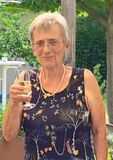 &Auml;ltere Frau mit Brille h&auml;lt ein Sektglas in der Hand, tr&auml;gt ein blumiges Oberteil und steht in einem bepflanzten Garten.