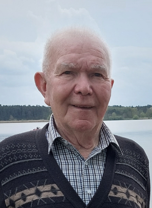 &Auml;lterer Mann mit grauen Haaren, im karierten Hemd und gemustertem Pullover, steht am Ufer eines Sees.