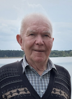 &Auml;lterer Mann mit grauen Haaren, im karierten Hemd und gemustertem Pullover, steht am Ufer eines Sees.