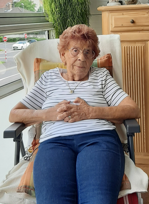 &Auml;ltere Frau mit rotem Haar in Streifen-T-Shirt sitzt entspannt in einem Sessel, H&auml;nde auf dem Bauch gefaltet.