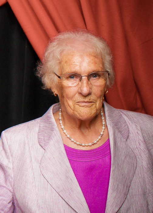 Portr&auml;t einer &auml;lteren Frau mit Brille, in einem grauen Blazer und einer Perlenkette, vor einem rotem Hintergrund.