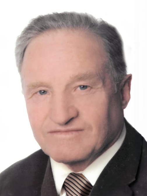 Portr&auml;t eines &auml;lteren Mannes mit grauen Haaren, in einem schwarzen Anzug und gestreifter Krawatte vor hellem Hintergrund.