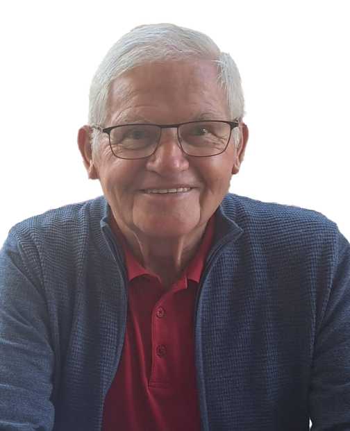 L&auml;chelnder &auml;lterer Mann mit Brille, rotem Polohemd und blauer Strickjacke vor einem wei&szlig;en Hintergrund.