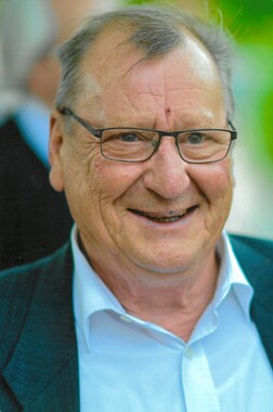 L&auml;chelnder &auml;lterer Mann mit Brille und Anzug, vor unscharfem Hintergrund.