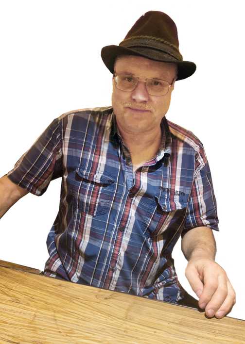 Mann mit Brille und Feldhut sitzt an einem Holztisch, tr&auml;gt ein kariertes Hemd und schaut in die Kamera.