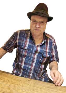 Mann mit Brille und Feldhut sitzt an einem Holztisch, tr&auml;gt ein kariertes Hemd und schaut in die Kamera.