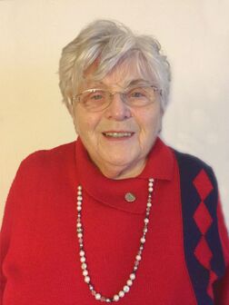 &Auml;ltere Frau mit grauen Haaren, Brille und rotem Pullover, l&auml;chelt vor einer hellen Wand. Sie tr&auml;gt eine Perlenkette.