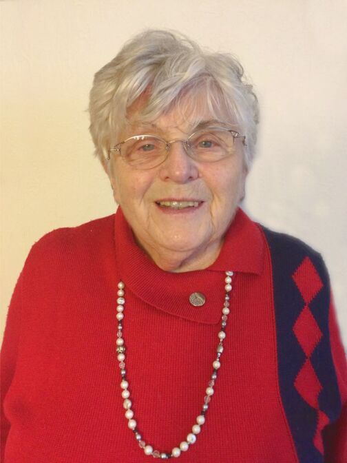 &Auml;ltere Frau mit grauen Haaren, Brille und rotem Pullover, l&auml;chelt vor einer hellen Wand. Sie tr&auml;gt eine Perlenkette.