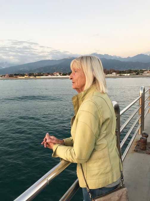 &Auml;ltere Frau mit blonden Haaren schaut nachdenklich &uuml;ber eine Holzbr&uuml;cke auf ruhiges Wasser und Berge im Hintergrund.