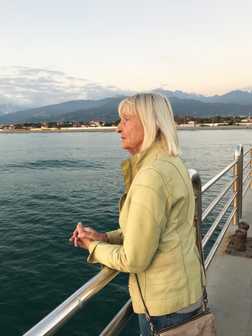 &Auml;ltere Frau mit blonden Haaren schaut nachdenklich &uuml;ber eine Holzbr&uuml;cke auf ruhiges Wasser und Berge im Hintergrund.