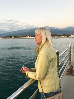 &Auml;ltere Frau mit blonden Haaren schaut nachdenklich &uuml;ber eine Holzbr&uuml;cke auf ruhiges Wasser und Berge im Hintergrund.