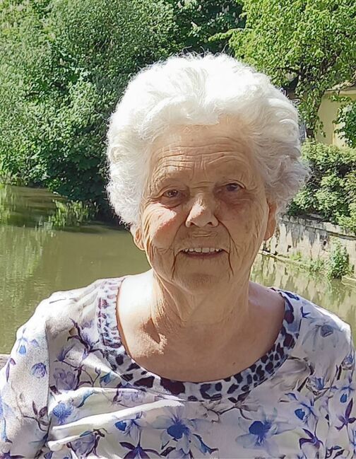 &Auml;ltere Frau mit kurzem, grauem Haar, l&auml;chelt vor einem Gew&auml;sser und gr&uuml;ner Umgebungsvegetation. Sie tr&auml;gt ein blumiges Oberteil.