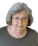 Ältere Frau mit Brille und grauen Haaren, trägt eine Perlenkette und ein dunkles Oberteil, lächelt in die Kamera.