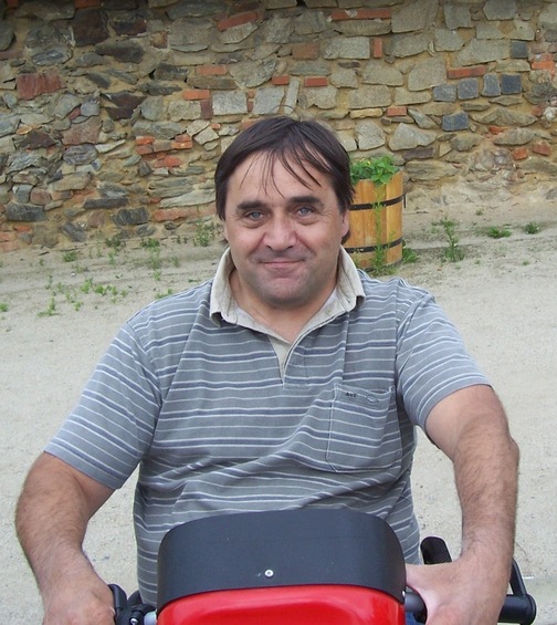 Mann sitzt auf einem roten Fahrzeug vor einer steinernen Wand in einer l&auml;ndlichen Umgebung.