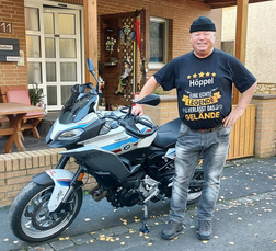 Mann in schwarzem T-Shirt steht neben einer BMW-Motorrad auf einem Gehweg vor einem Haus mit Ziegelmauer.