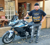 Mann in schwarzem T-Shirt steht neben einer BMW-Motorrad auf einem Gehweg vor einem Haus mit Ziegelmauer.