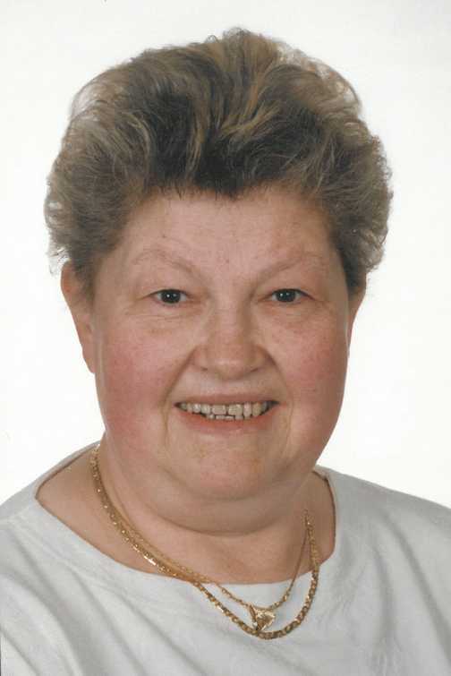 Portr&auml;t einer &auml;lteren Frau mit kurzem, lockigem Haar und Goldschmuck, l&auml;chelnd vor einem neutralen Hintergrund.