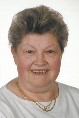 Portr&auml;t einer &auml;lteren Frau mit kurzem, lockigem Haar und Goldschmuck, l&auml;chelnd vor einem neutralen Hintergrund.