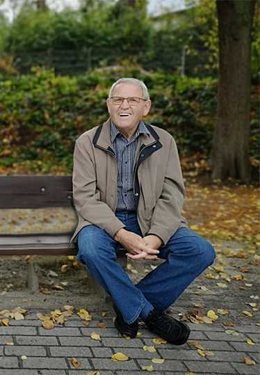 L&auml;chelnder &auml;lterer Mann sitzt entspannt auf einer Bank in einem herbstlichen Park mit buntem Laub.