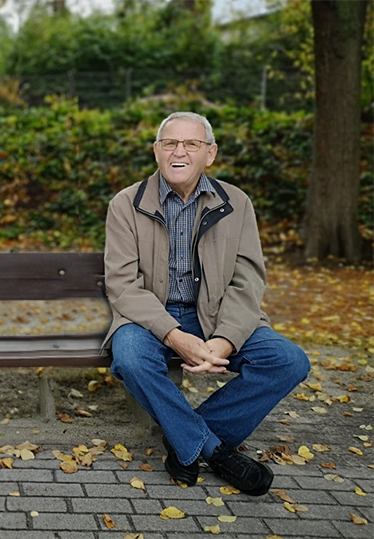 L&auml;chelnder &auml;lterer Mann sitzt entspannt auf einer Bank in einem herbstlichen Park mit buntem Laub.