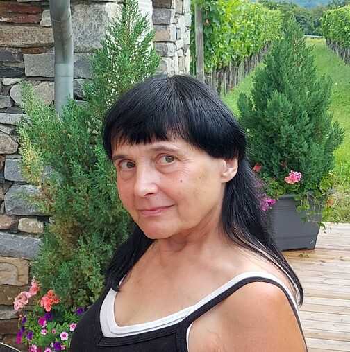 Portr&auml;t einer Frau mit schwarzem Haar, die vor einer Steinmauer und gr&uuml;nen Pflanzen steht. Bl&uuml;hende Blumen sind sichtbar.