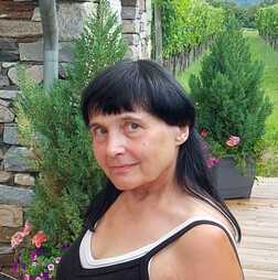Portr&auml;t einer Frau mit schwarzem Haar, die vor einer Steinmauer und gr&uuml;nen Pflanzen steht. Bl&uuml;hende Blumen sind sichtbar.