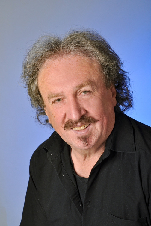 Portrait eines l&auml;chelnden Mannes mit grauen Haaren, in schwarzer Kleidung vor einem hellblauen Hintergrund.