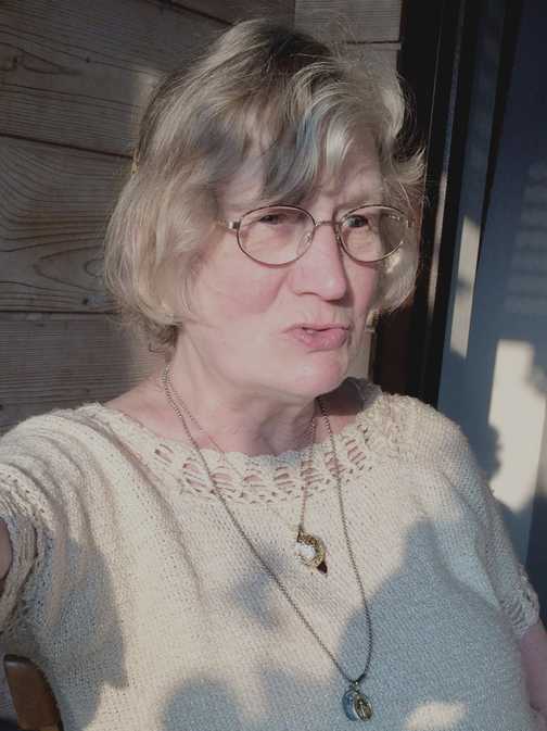 Portr&auml;t einer &auml;lteren Frau mit blonden, gewellten Haaren, Brille und Schmuck, vor einer Holzverkleidung im Sonnenlicht.