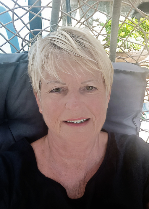 Portrait einer Frau mit kurzen, blonden Haaren, die in einem geflochtenen H&auml;ngesessel sitzt und l&auml;chelt.