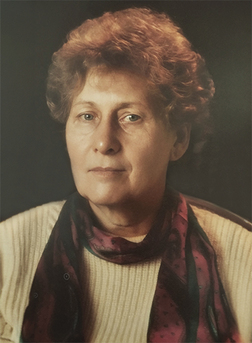 Portr&auml;t einer &auml;lteren Frau mit lockigem Haar, einem hellen Pullover und einem gepunkteten Schal. Der Hintergrund ist dunkel.